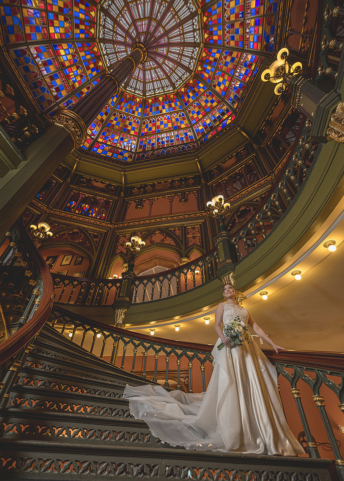 Bride holding a bouquet on the cast-iron spiral staircase beneath the stained-glass dome of the Old State Capitol in Baton Rouge