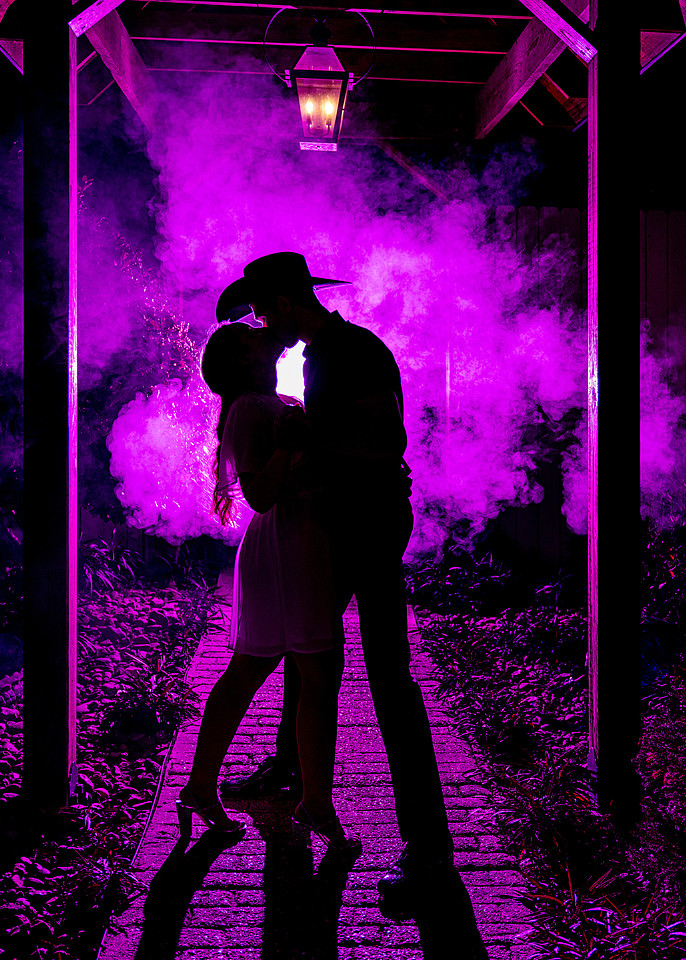 Silhouette of an engaged couple kissing beneath a lantern-lit walkway, the man in a cowboy hat, backlit through clouds of magenta smoke